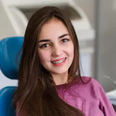 Uśmiechnięta dziewczyna z brązowymi włosami siedzi na niebieskim fotelu dentystycznym, prezentując swoje różowe ligatury na aparacie ortodontycznym.