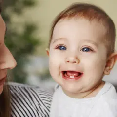 Uśmiechnięte dziecko z jednym ząbkiem, mama patrzy na nie z czułością. To początek kolejności wychodzenia zębów.