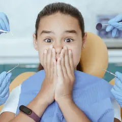 Przerażona dziewczynka w gabinecie dentystycznym, otoczona przez ręce w rękawiczkach z narzędziami. Czy to objawy wyrzynania szóstek?