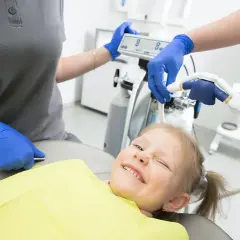 Uśmiechnięta dziewczynka u dentysty, która nie boi się, czy zęby 5 wypadają. Personel medyczny przygotowuje sprzęt.