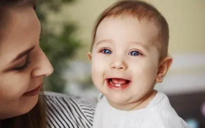 Uśmiechnięte dziecko z jednym ząbkiem, mama patrzy na nie z czułością. To początek kolejności wychodzenia zębów.