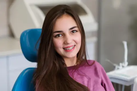 Uśmiechnięta dziewczyna z brązowymi włosami siedzi na niebieskim fotelu dentystycznym, prezentując swoje różowe ligatury na aparacie ortodontycznym.