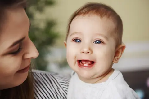 Uśmiechnięte dziecko z jednym ząbkiem, mama patrzy na nie z czułością. To początek kolejności wychodzenia zębów.
