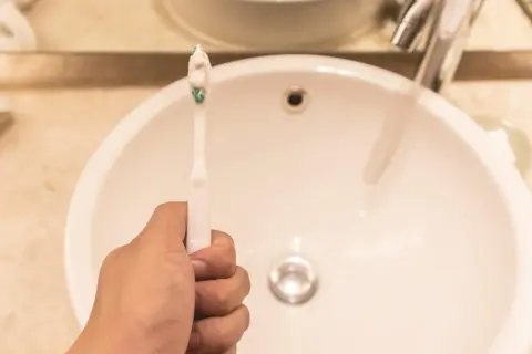 Ręka trzymająca szczoteczkę do zębów z pastą nad umywalką. Czy to mycie zębów przed czy po śniadaniu?