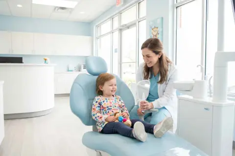 Uśmiechnięta dziewczynka na fotelu dentystycznym, trzymająca pluszaka. Dentystka pokazuje jej lusterko. To ich pierwsza wizyta u dentysty, ale oboje są zrelaksowani.
