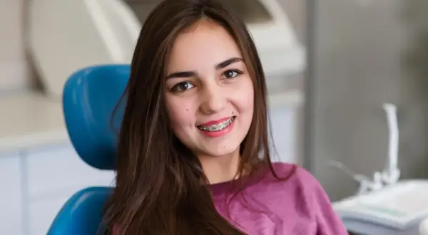 Uśmiechnięta dziewczyna z brązowymi włosami siedzi na niebieskim fotelu dentystycznym, prezentując swoje różowe ligatury na aparacie ortodontycznym.