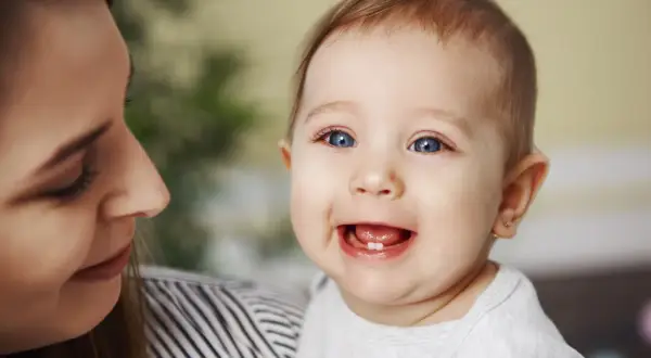 Uśmiechnięte dziecko z jednym ząbkiem, mama patrzy na nie z czułością. To początek kolejności wychodzenia zębów.