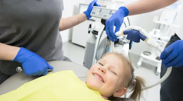 Uśmiechnięta dziewczynka u dentysty, która nie boi się, czy zęby 5 wypadają. Personel medyczny przygotowuje sprzęt.