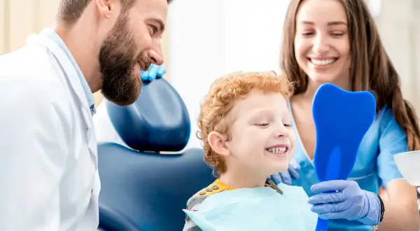 Uśmiechnięty chłopiec po fluoryzacji zębów ogląda swoje zdrowe ząbki w lusterku w kształcie zęba, zadowolony z wizyty u dentysty.