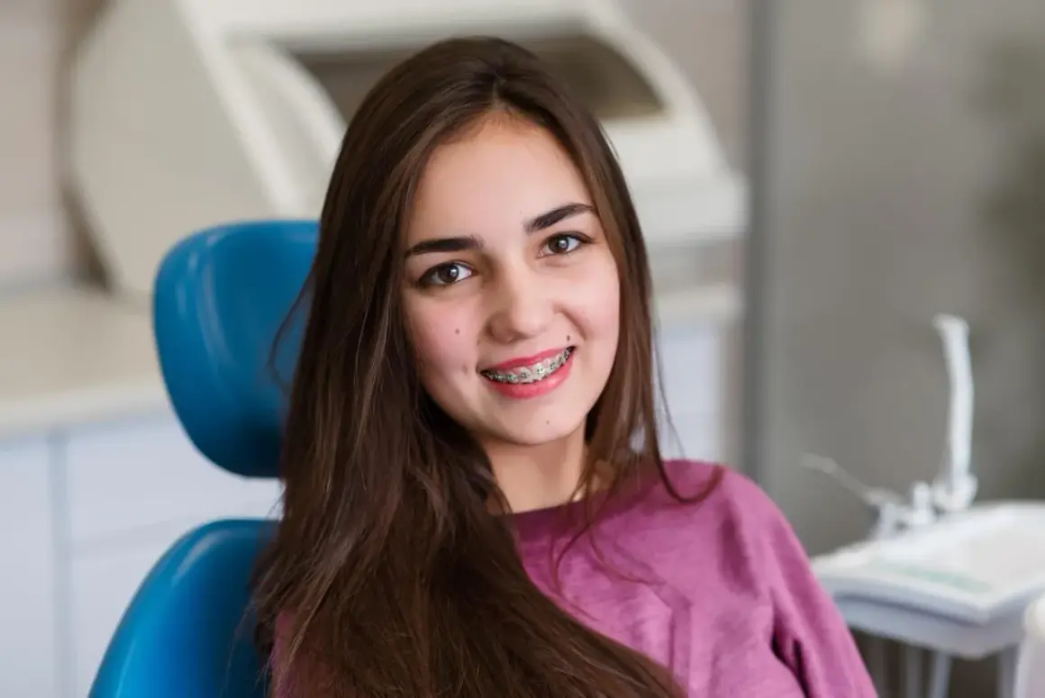 Uśmiechnięta dziewczyna z brązowymi włosami siedzi na niebieskim fotelu dentystycznym, prezentując swoje różowe ligatury na aparacie ortodontycznym.