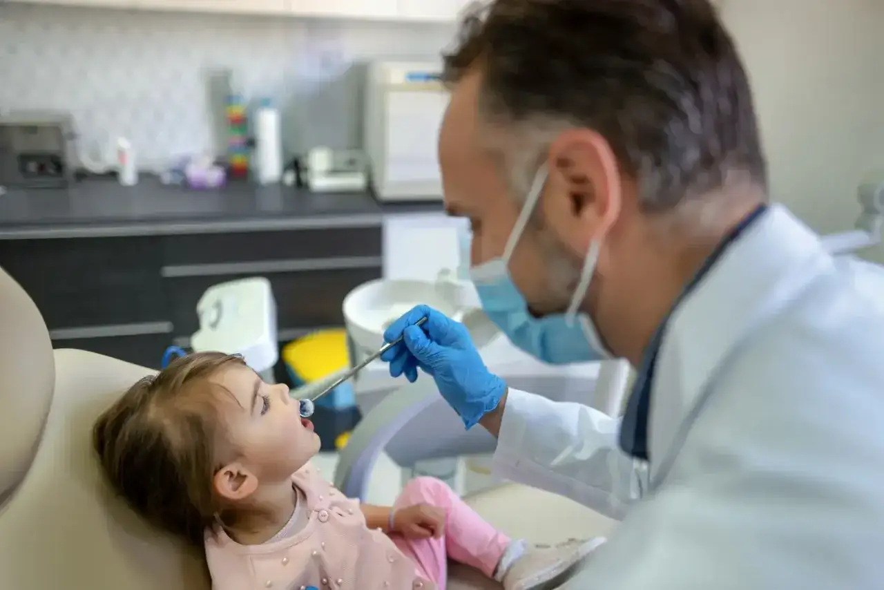Mała dziewczynka ma swoją pierwszą wizytę u dentysty. Lekarz w masce i rękawiczkach bada jej ząbki.