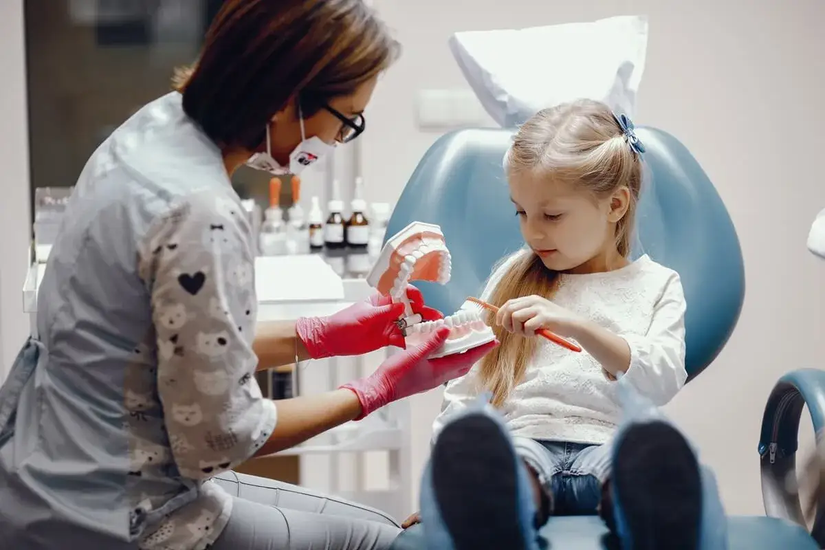 Wizyta u dentysty: mała dziewczynka uczy się szczotkować zęby na modelu szczęki, asystowana przez higienistkę.