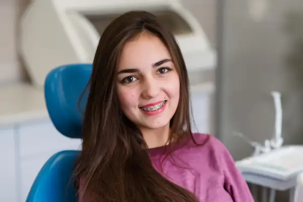 Uśmiechnięta dziewczyna z brązowymi włosami siedzi na niebieskim fotelu dentystycznym, prezentując swoje różowe ligatury na aparacie ortodontycznym.