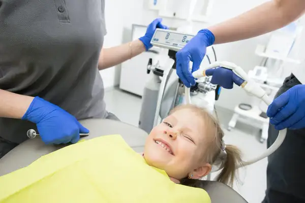 Uśmiechnięta dziewczynka u dentysty, która nie boi się, czy zęby 5 wypadają. Personel medyczny przygotowuje sprzęt.