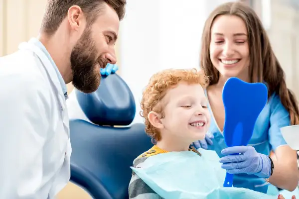 Uśmiechnięty chłopiec po fluoryzacji zębów ogląda swoje zdrowe ząbki w lusterku w kształcie zęba, zadowolony z wizyty u dentysty.
